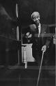 A black and white photograph of a man in a dark suit, holding a fedora in one hand and a cane i…